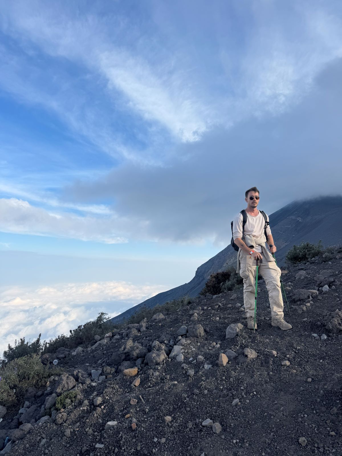 Person standing on a mountain peak with a backpack, surrounded by clouds and blue sky.