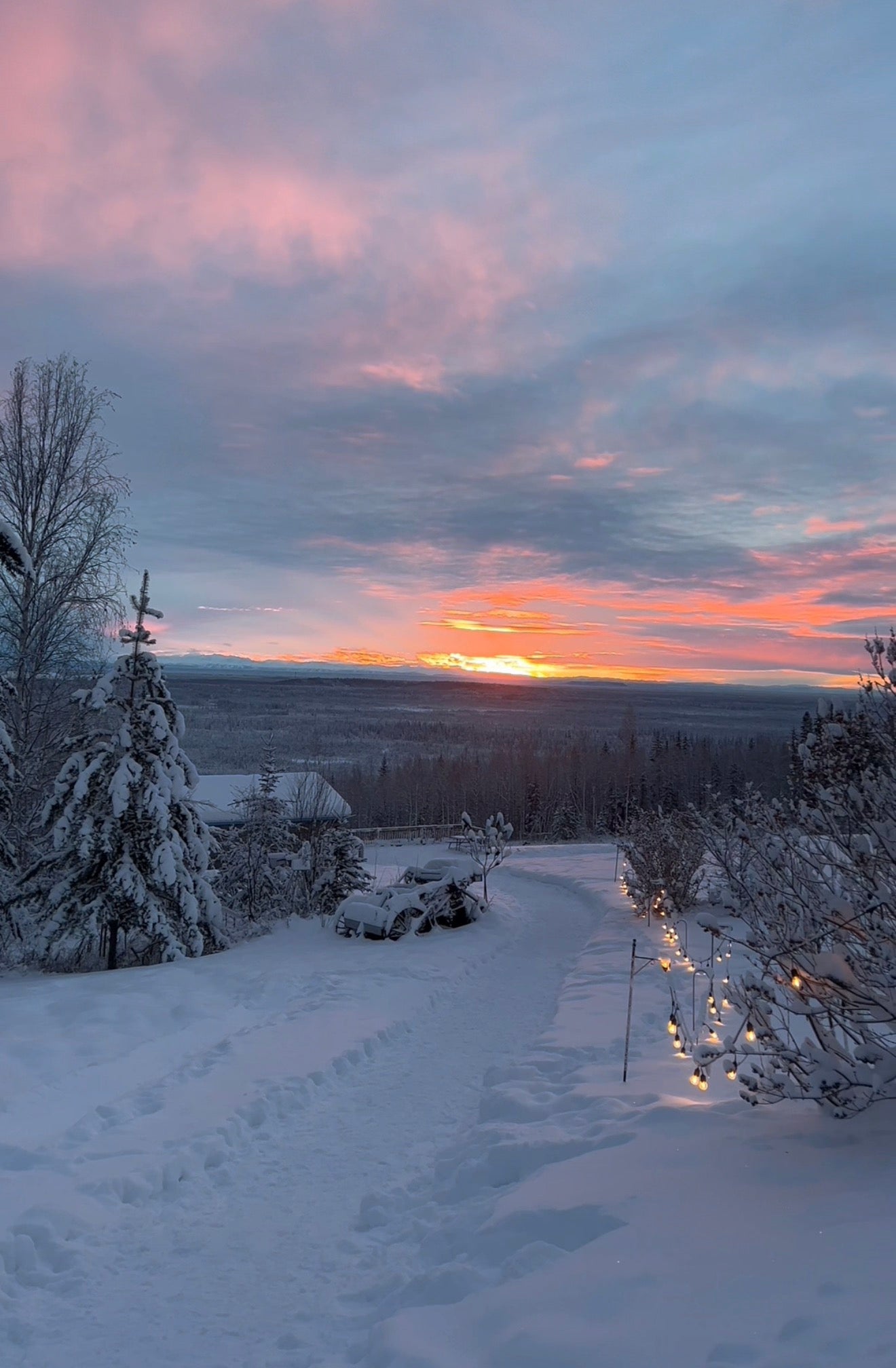 Snowy landscape with a sunset over a mountain range, trees, and a path.