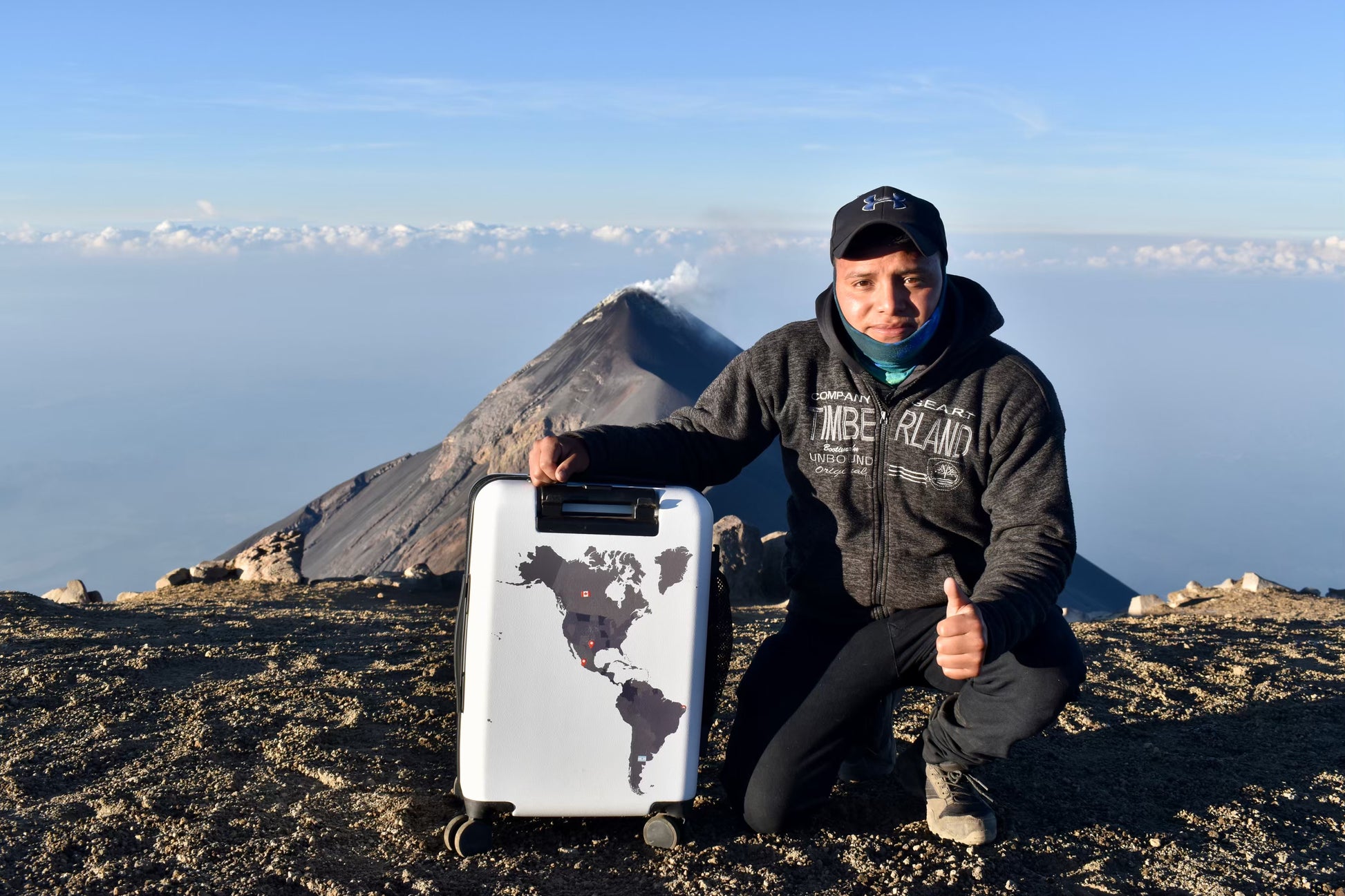 Person on a mountain peak holding a trvld white carry on suitcase with world map design, wearing a dark jacket and cap.